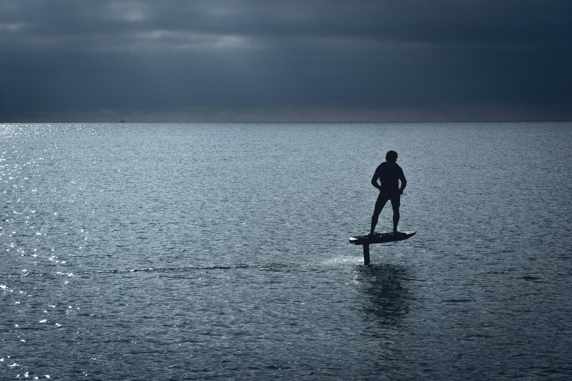 A silhouetted e-foiler standing above calm water under an overcast sky